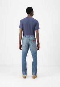 Jeans azules con corte recto y diseño de bolsillo trasero, combinados con una camiseta de color marino. El modelo está de espaldas, mostrando el conjunto.