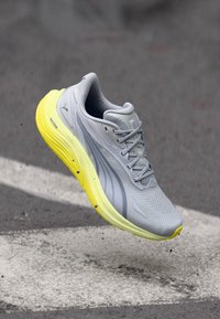 Γκρι και κίτρινο αθλητικό παπούτσι στον αέρα πάνω από άσφαλτο με λευκές οδικές σημάνσεις, που δείχνει λεπτομερή σχεδίαση και υφασμένο πέλμα.