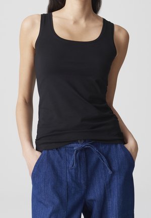Kvinde iført en sort ærmeløs t-shirt med rund hals og blå bukser med snøre, hænderne hvilende i bukselommerne, på lysegrå baggrund.