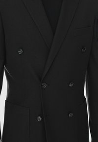 Dwurzędowa czarna marynarka wykonana z teksturowanej tkaniny. Posiada czarne guziki, klapy w kształcie litery V oraz jedną kieszeń na piersi po lewej stronie.