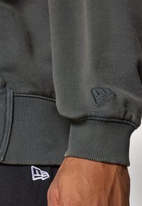 Tessuto di felpa verde scuro con polsini a costine, caratterizzato da un ricamo discreto del logo New Era in tonalità contrastanti sulla manica e sul punto vita.