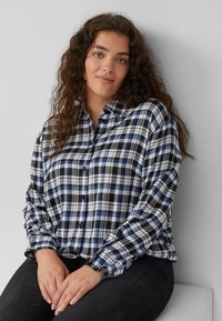 Frau mit lockigem braunem Haar, die ein blaues, schwarzes und weißes kariertes Hemd mit Knöpfen trägt und an einer schlichten hellgrauen Wand sitzt.