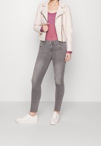 Vrouw gekleed in een lichtroze bikerjack, mauve langemouwen top, grijze skinny jeans en witte sneakers, staand tegen een effen achtergrond.