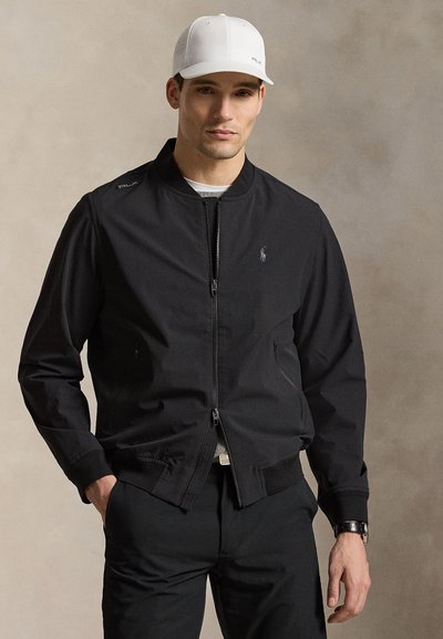 Homme portant une veste noire zippée, un pantalon noir, une casquette blanche et une montre-bracelet, debout devant un fond neutre uni.
