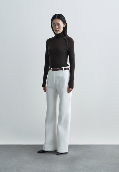 Top cu guler înalt de culoare maro, asortat cu pantaloni albi cu talie înaltă. Pantalonii au buzunare frontale, iar un curea maro accentuează talia.