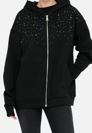 Zwarte hoodie met rits met zilverkleurige studs en strass-steentjes op de bovenste voorkant, gedragen door een persoon met één hand in de zak en zwarte broek.