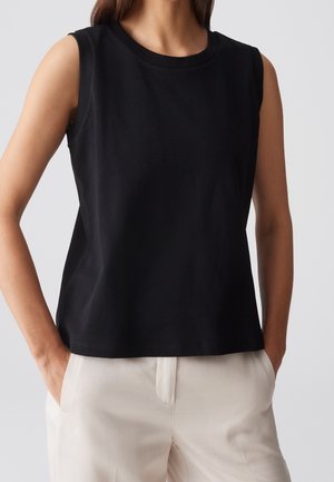 Femme portant un haut noir sans manches et un pantalon clair, debout les mains dans les poches devant un fond uni.