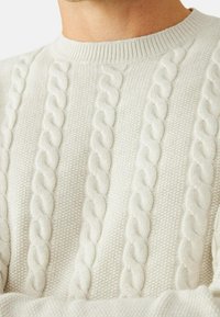 Pull en maille couleur crème présentant des motifs torsadés le long de lignes verticales, avec un col rond côtelé et un tissu texturé.