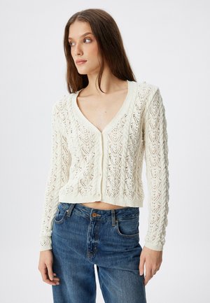 Cardigan en tricot crème à manches courtes avec un motif ajouré sophistiqué et des accents de perles. Présente un décolleté en V profond et une coupe cropped.