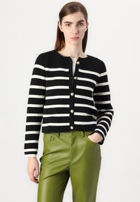 Cardigan a righe bianche e nere, lavorato a maglia, con colletto rotondo e bottoni dorati, indossato sopra pantaloni in pelle verde. Presenta due tasche frontali.