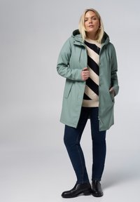 Mintgroene, waterdichte jas met een capuchon, voorzien van een voorrits en drukknoopsluiting. Draagt over een gestreepte trui en donkere jeans, met zwarte laarzen.