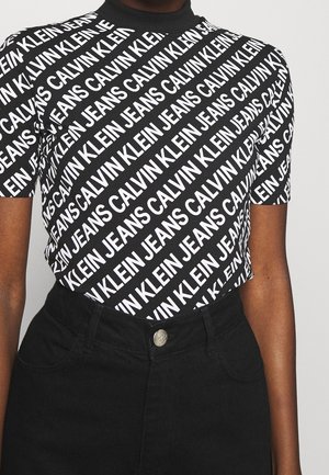 Personne portant un t-shirt noir à manches courtes avec un texte blanc répété "Calvin Klein Jeans" et un pantalon noir taille haute avec un bouton.