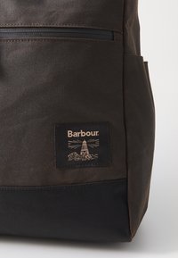 Dark brown and black Barbour backpack with front zip pocket and a lighthouse logo patch on the lower front panel.