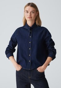 Camicia in denim scuro con colletto alla mandarino, chiusura con bottoni e maniche arrotolate. Cuciture a contrasto visibili e tasche laterali.