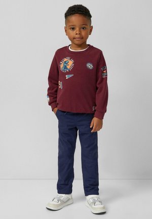 Bordeauxfarbener Pullover mit grafischen Designs, marineblaue Hose und weiße Sneakers. Merkmale sind gerippte Bündchen und mehrere bunte Patches.