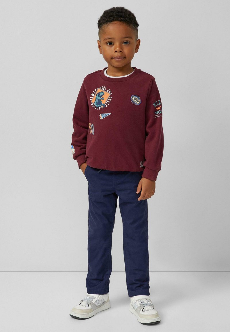 Sweatshirt marron avec des graphismes, pantalon bleu marine et baskets blanches. Caractéristiques : poignets côtelés et plusieurs patchs colorés.