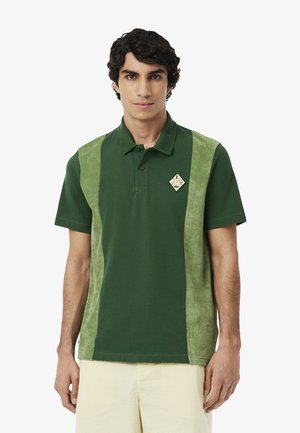 Polo verde con paneles laterales texturizados, que presenta un parche en forma de diamante en el pecho izquierdo y un cuello clásico.