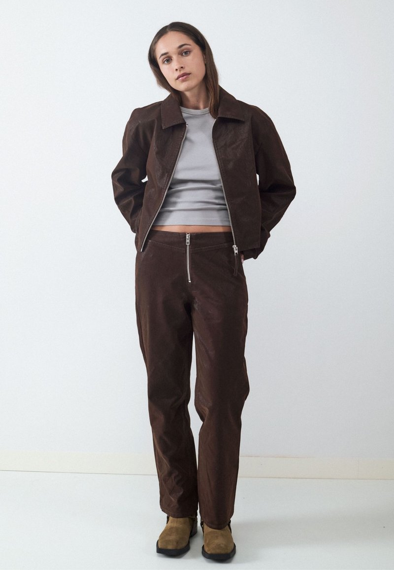 Young woman wearing brown jacket and pants with front zippers, gray cropped shirt, and tan boots, standing against a white wall.