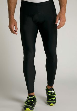 Schwarze Radsportleggins mit einer engen Passform, ausgestattet mit einem kreisförmigen Polsterdesign an der Vorderseite, kombiniert mit schwarzen Sportschuhen mit grünen Akzenten.
