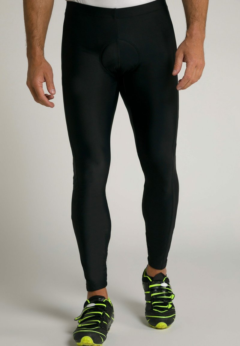 Schwarze Radsportleggins mit einer engen Passform, ausgestattet mit einem kreisförmigen Polsterdesign an der Vorderseite, kombiniert mit schwarzen Sportschuhen mit grünen Akzenten.