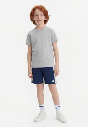 Rødharret dreng står, iført en grå kortærmet t-shirt, marineblå sportsshorts med hvide sider, hvide sokker og hvide sneakers.