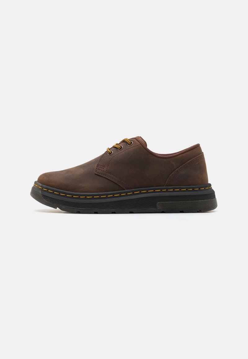 Chaussure basse en cuir marron avec semelle épaisse noire et coutures jaunes, comportant trois œillets avec lacets marron et jaunes, vue de côté.