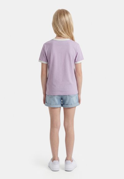 Enfant aux longs cheveux blonds se tenant de dos, portant un t-shirt lavande à manches courtes, un short en jean clair et des baskets blanches sur fond blanc.