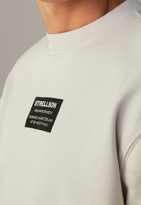 Ljusgrå sweatshirt med en texturerad tyg, som har en svart rektangulär etikett med vit text på vänster sida. Ribbad detalj vid halsringningen.
