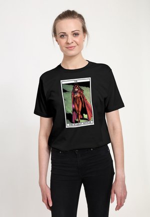 Ung kvinde iført sort t-shirt med tegneseriestil Scarlet Witch-figur i rød dragt og kappe, stående selvsikkert.