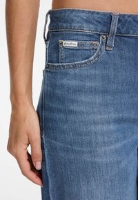 Mėlynos džinsinės kelnės su išblukusiu tonu. Turintis priekinį kišeni, matomą siuvimą ir etiketę su užrašu "Guess Jeans."