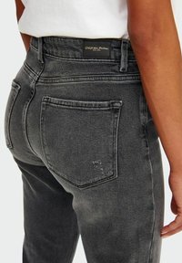 Jeans de mezclilla negros desgastados con cintura alta, cinco bolsillos y una etiqueta de marca. La tela presenta un ligero deterioro cerca del bolsillo.