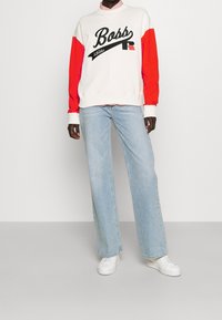 Felpa bianca con maniche rosse, logo "Boss" nero, abbinata a jeans a gamba larga blu chiaro e sneakers bianche.