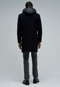 Abrigo de lana negro con capucha acolchada y desmontable. Presenta un diseño ajustado, bolsillos laterales con abertura y un corte largo, combinado con jeans grises y zapatos negros.