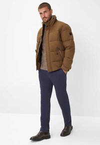 Braune Daunenjacke mit hohem Kragen, zwei Seitentaschen und Reißverschluss, kombiniert mit einem leichten Strickpullover und navyblauen Hosen. Braune Knöchelstiefel.