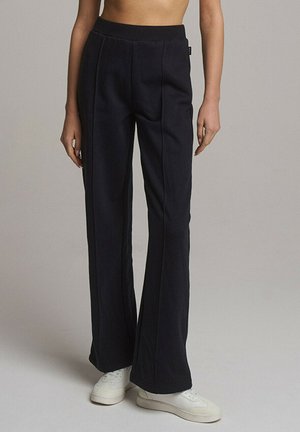 Czarne spodnie flare o gładkiej fakturze, wysoki stan i fałdy z przodu. Posiadają elastyczny pas i są stylizowane z białymi sneakersami.