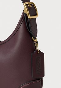 Primo piano di una tracolla in pelle bordeaux scuro con una fibbia dorata e un'etichetta in pelle abbinata embossata con "Coach Leatherware".