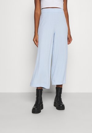 Vrouw die lichtblauwe wijde cropped broek, wit cropped top en zwarte veterplateau laarzen draagt, staand tegen een witte achtergrond.