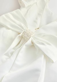 Witte jurk met een gladde satijnen afwerking, voorzien van een grote strik op de taille versierd met een cluster kleine parels en een gouden accent.