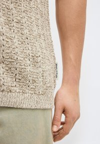 Maglione beige in maglia testurizzata con etichetta Marc O'Polo indossato sopra pantaloni verde smeraldo tenue, che mostra la mano destra rilassata e l'avambraccio di una persona.