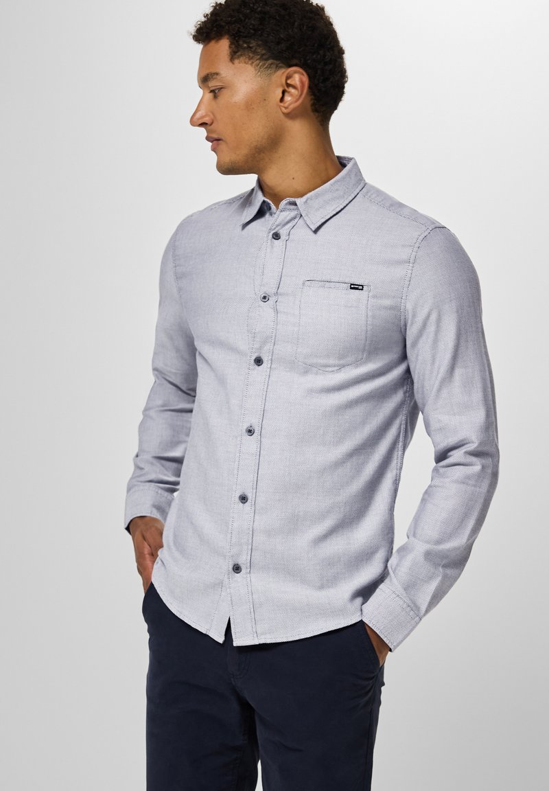 Camicia azzurro chiaro con maniche lunghe, taschino sul petto e un sottile motivo testurizzato. Rifinita con bottoni scuri.