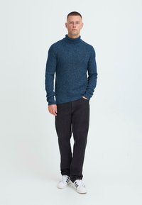 Temno modra pletena pulover turtleneck z teksturirano površino, v kombinaciji s črnimi hlačami in belimi supergi, ki imajo črne črte.