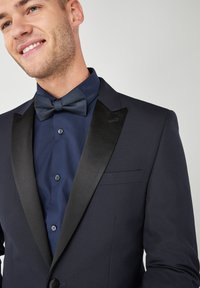 Giacca da smoking navy con rever in satin nero, indossata sopra una camicia blu scuro, con un papillon strutturato e dettagli dei bottoni.