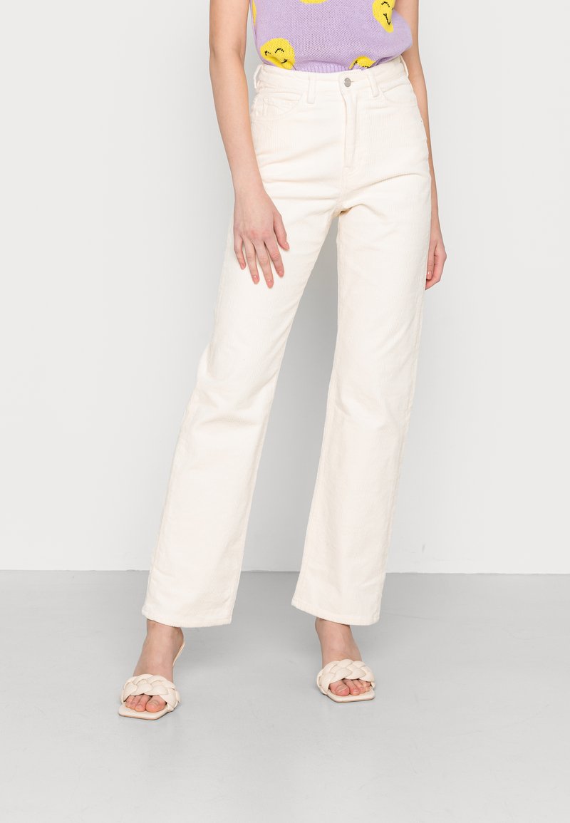 Calças largas de veludo branco com bolsos frontais, cintura alta e textura subtil, combinadas com sandálias de tira trançada creme e uma blusa roxa.