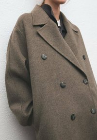 Dubbelgeknoopte bruine wollen jas met grote zwarte knopen, brede revers en een oversized pasvorm. Zachte textuur met een gestructureerd ontwerp.