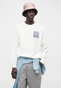 Άνδρας που φοράει ροζ καπέλο τύπου bucket, λευκό μακρυμάνικο πουκάμισο με σχέδιο, μπλε φούτερ δεμένο στη μέση και γκρι ριγέ παντελόνι, μπροστά από λευκό φόντο.