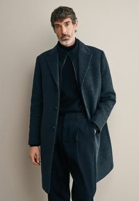Abrigo de lana gris oscuro con solapas con muesca, botones dobles y bolsillos en ángulo, superpuesto a un suéter negro de canalé y pantalones a medida.