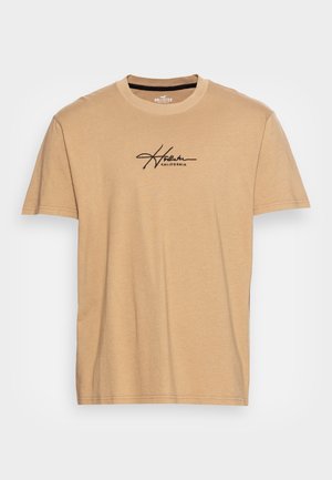 Beżowy t-shirt z krótkim rękawem z czarnym logo "Hollister California" w stylu pisma, umieszczonym centralnie na piersiach, okrągły dekolt.