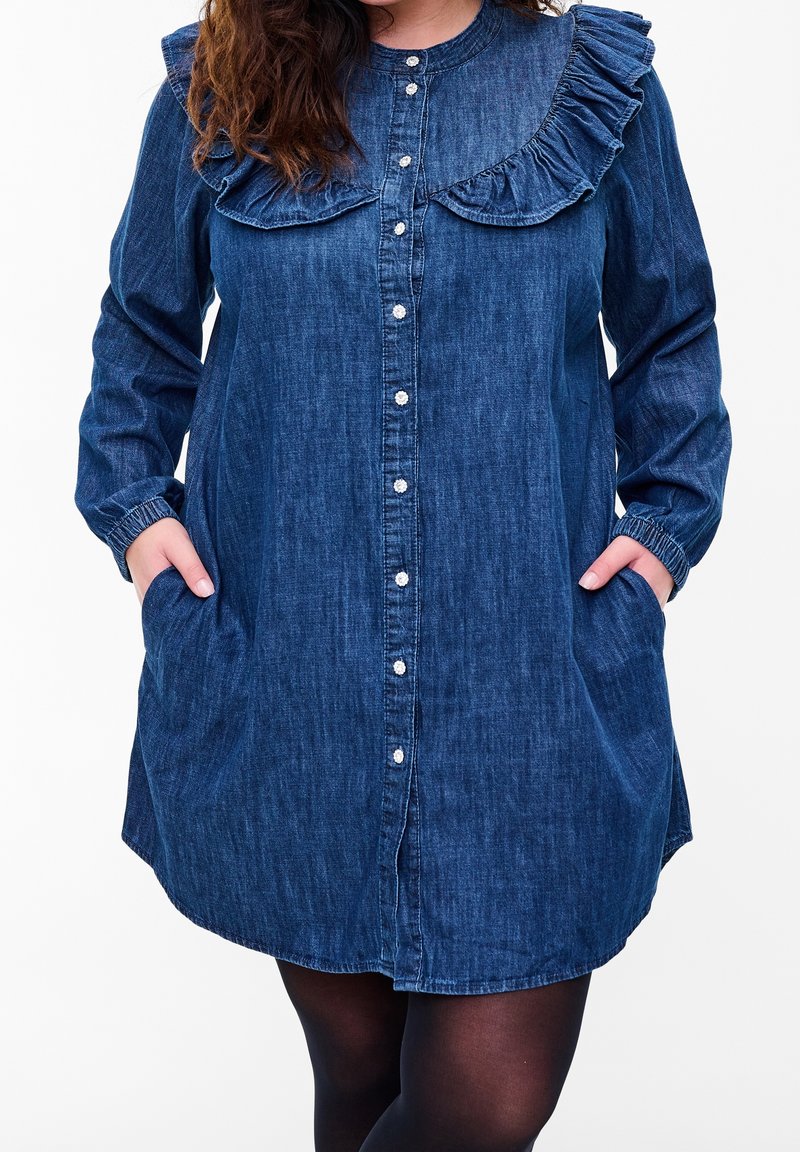 Femme portant une robe en jean bleu à manches longues avec un col à volants, des boutons sur le devant et les mains dans les poches, vue du cou jusqu'à la cuisse.