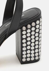 Sandale noire à talon haut avec un talon bloc épais décoré de multiples clous circulaires argentés brillants, et une lanière noire à travers le pied.