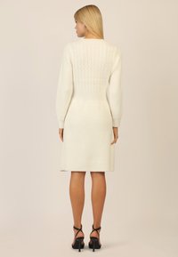 Weißes gestricktes Kleid mit gerippter Textur und Zopfmuster auf der Rückseite. Lange Ärmel, taillierte Passform und knielanger Saum.